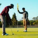Two golfers on a putting green, one preparing to putt while the other holds the flagstick.