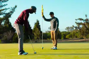 Two golfers on a putting green, one preparing to putt while the other holds the flagstick.