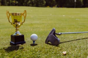 A golden trophy, golf ball on a tee, and a putter on a green golf course